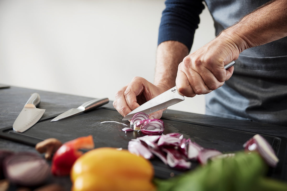 A person precisely cuts a red onion with the Fissler Essential® Utility Knife, next to characteristic fresh vegetables and clear design.