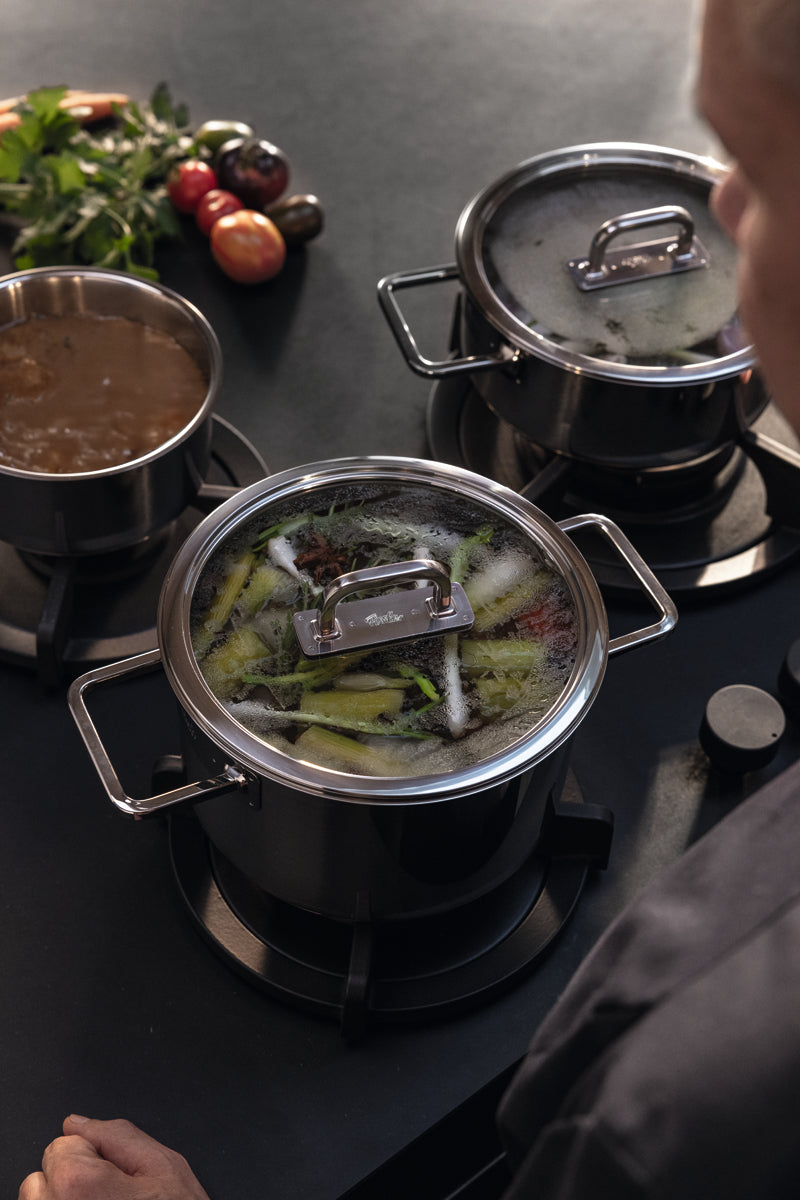 A person cooks with three high-quality Fissler stainless steel pots; broth, simmering vegetables, and fresh produce complete the scene.