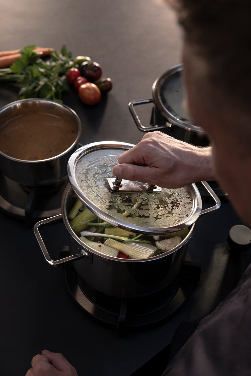 A person lifts the lid of a Fissler Viseo® Pro stainless steel pot filled with vegetables; the sauce is simmering and fresh vegetables are next to it.