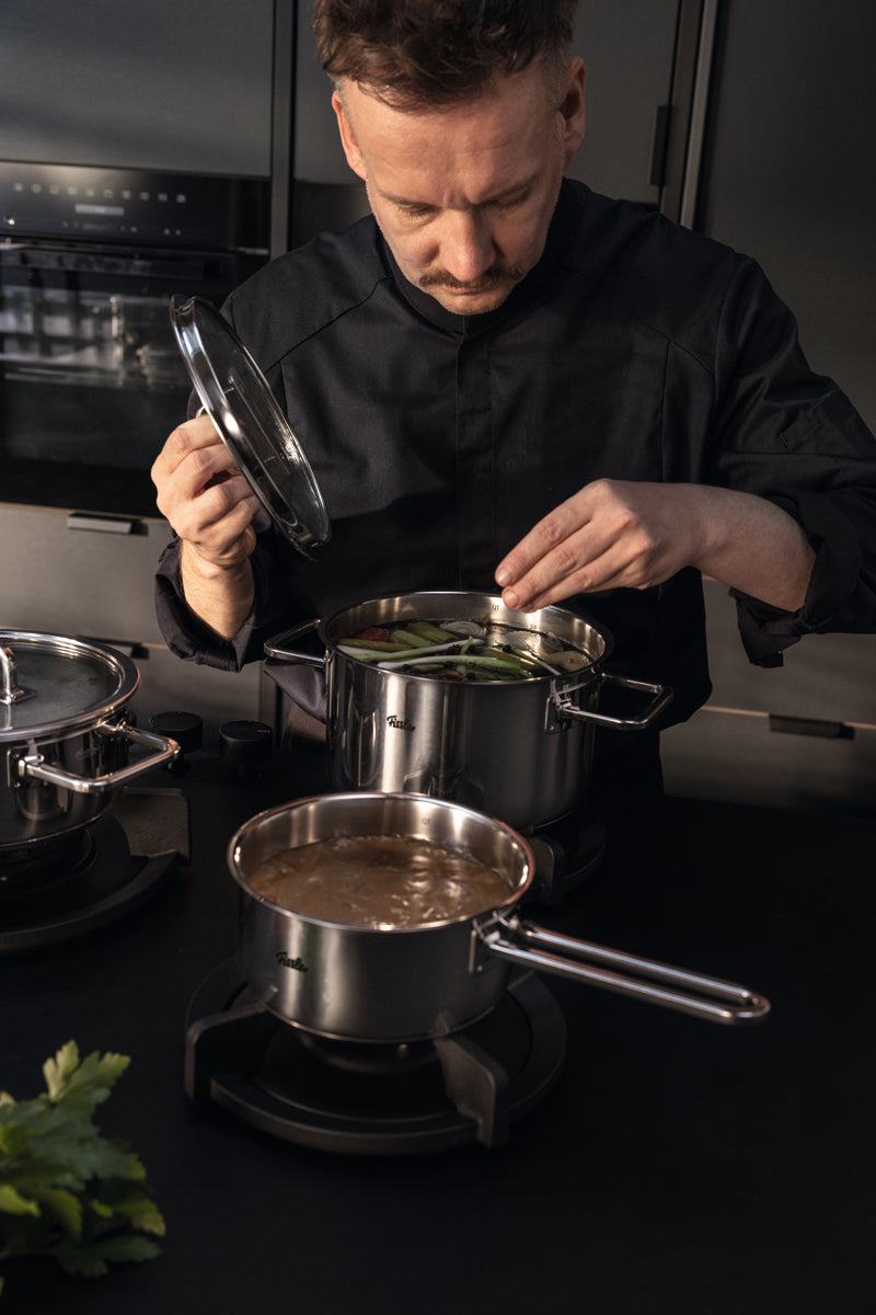 A chef puts green beans into a Fissler Viseo® Pro 16 cm saucepan; stainless steel pots simmer in an elegant, modern kitchen.