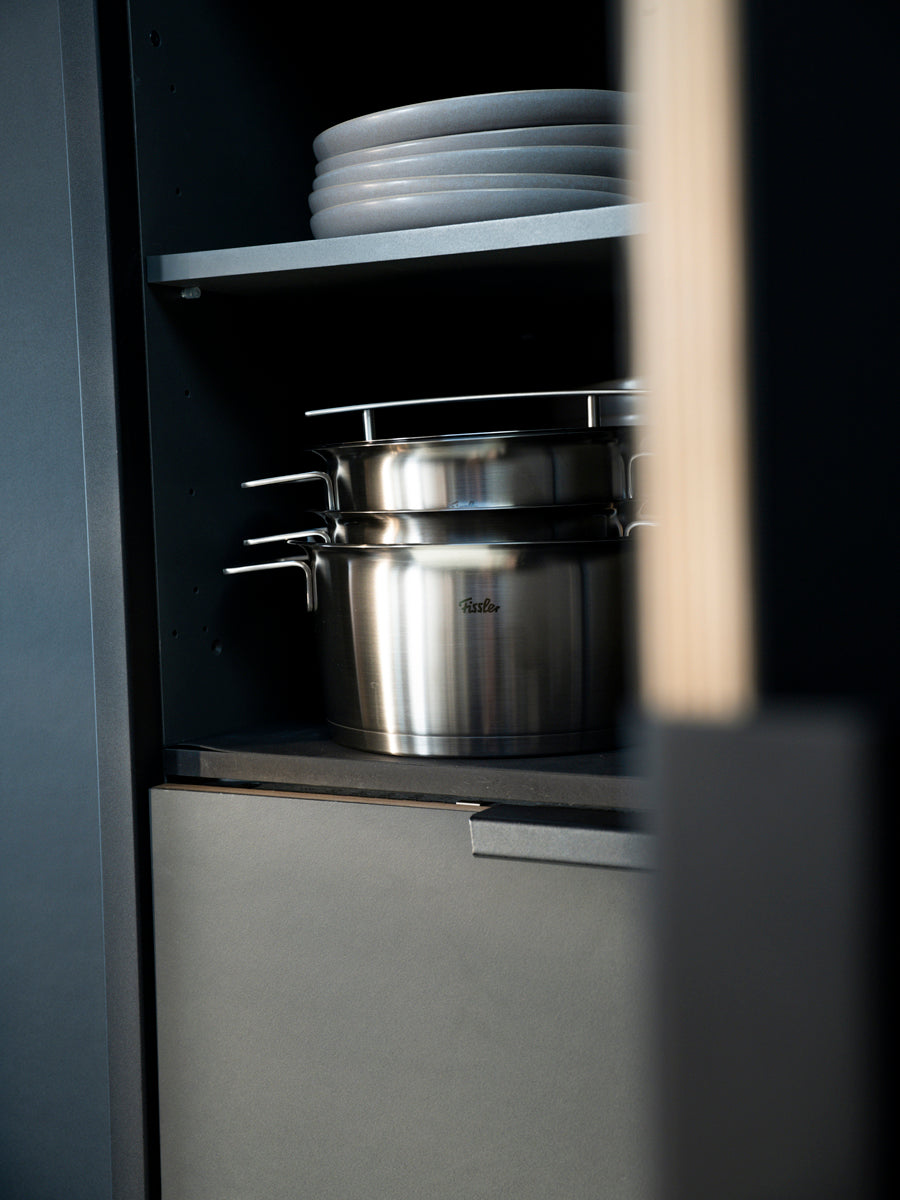 Über einem Topf aus der Fissler Phi Collection® sind elegante weiße Teller gestapelt, die in einem dunklen Küchenschrank das zeitlose Design aus deutscher Fertigung zur Geltung bringen.
