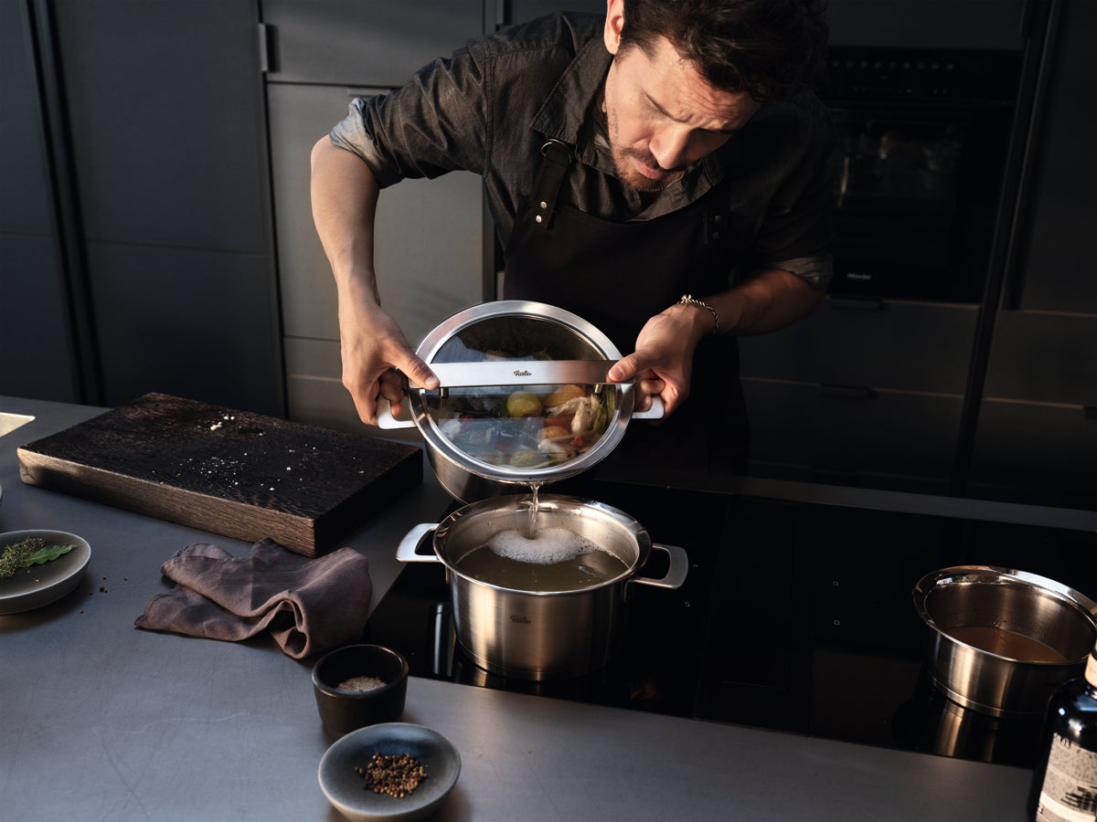 Ein Mann gibt Gemüse in einen Topf der Fissler Phi Collection® und demonstriert damit deutsche Präzision auf einer eleganten Küchenarbeitsplatte.