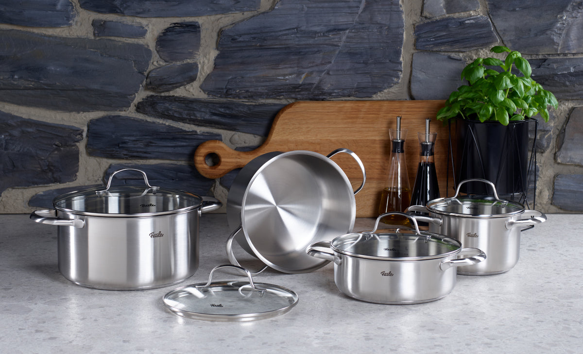 Erleben Sie Präzisionsingenieurkunst mit dem Fissler San Francisco Schmortopf-Set: drei hochwertige Edelstahltöpfe mit passenden Deckeln, hergestellt in Deutschland und elegant auf der Küchenarbeitsplatte arrangiert neben einem Holzschneidebrett, zwei Ölflaschen und einer farbenfrohen Basilikumpflanze im Topf. Vor dem Hintergrund einer edlen Steinwand besticht jeder Topf durch unseren charakteristischen „Superthermic-Boden“, der für eine hervorragende Wärmeverteilung und nahtlose Kompatibilität mit