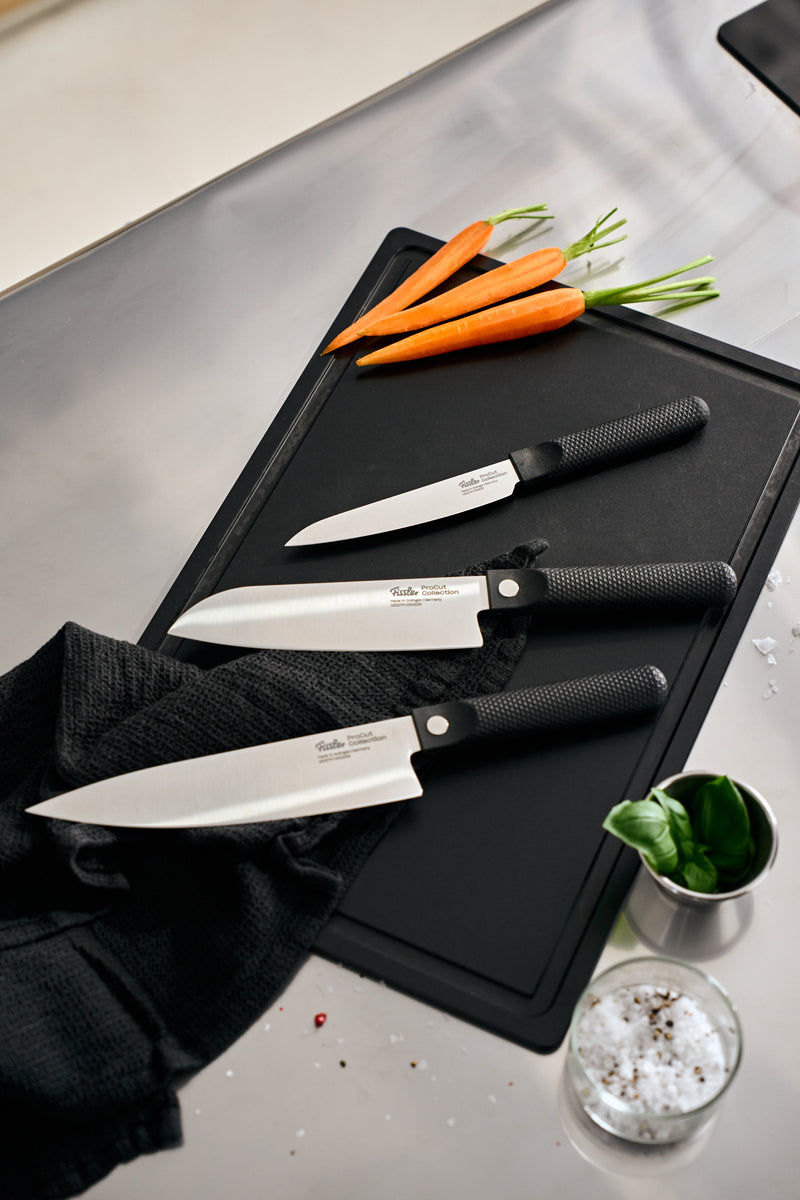 Experience Fissler precision and timeless design with the ProCut 3-piece knife set—Universalmesser, Santokumesser, and Kochmesser—expertly arranged on a sleek black cutting board. Showcased beside a sophisticated black towel, freshly cut carrots, vibrant basil leaves, and coarse salt in a small cup atop a pristine stainless steel countertop. Celebrating Fissler’s heritage of German engineering since 1845, these kitchen essentials embody durability and innovation for inspired daily cooking.