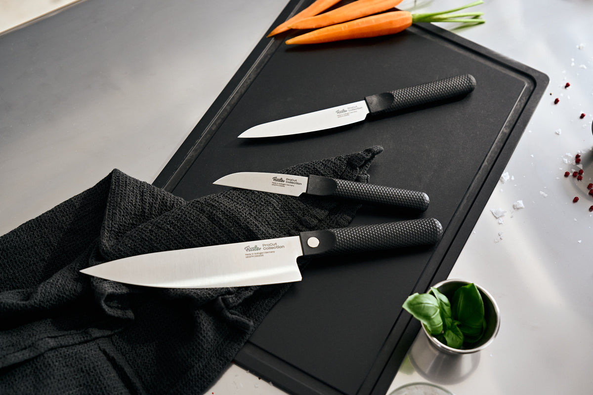 Experience the precision and timeless elegance of Fissler: Three expertly crafted kitchen knives from the Fissler ProCut 3-tlg. Set—a Gemüsemesser, Universalmesser, and Kochmesser—each with ergonomic black handles, lie on a sophisticated black cutting board. Fresh carrots, basil leaves, salt crystals, peppercorns, and a folded black cloth accentuate the scene atop a polished stainless steel countertop. Proudly Made in Germany since 1845—where innovation meets enduring quality.