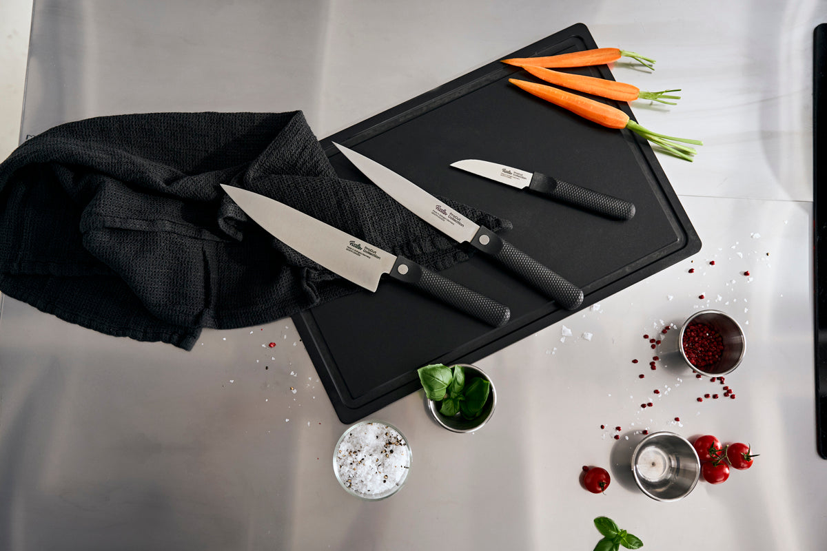 Experience the heritage of Fissler, founded in 1845 and renowned for precision-engineered, durable products “Made in Germany.” The Fissler ProCut 3-piece set—comprising the Gemüsemesser, Fleischmesser, and Kochmesser—is beautifully presented on a sleek black cutting board alongside a black towel. Accentuating the timeless design are fresh carrots, fragrant basil leaves, a bowl of salt, vibrant red peppercorns, and a candle—all arranged on an elegant marble surface.