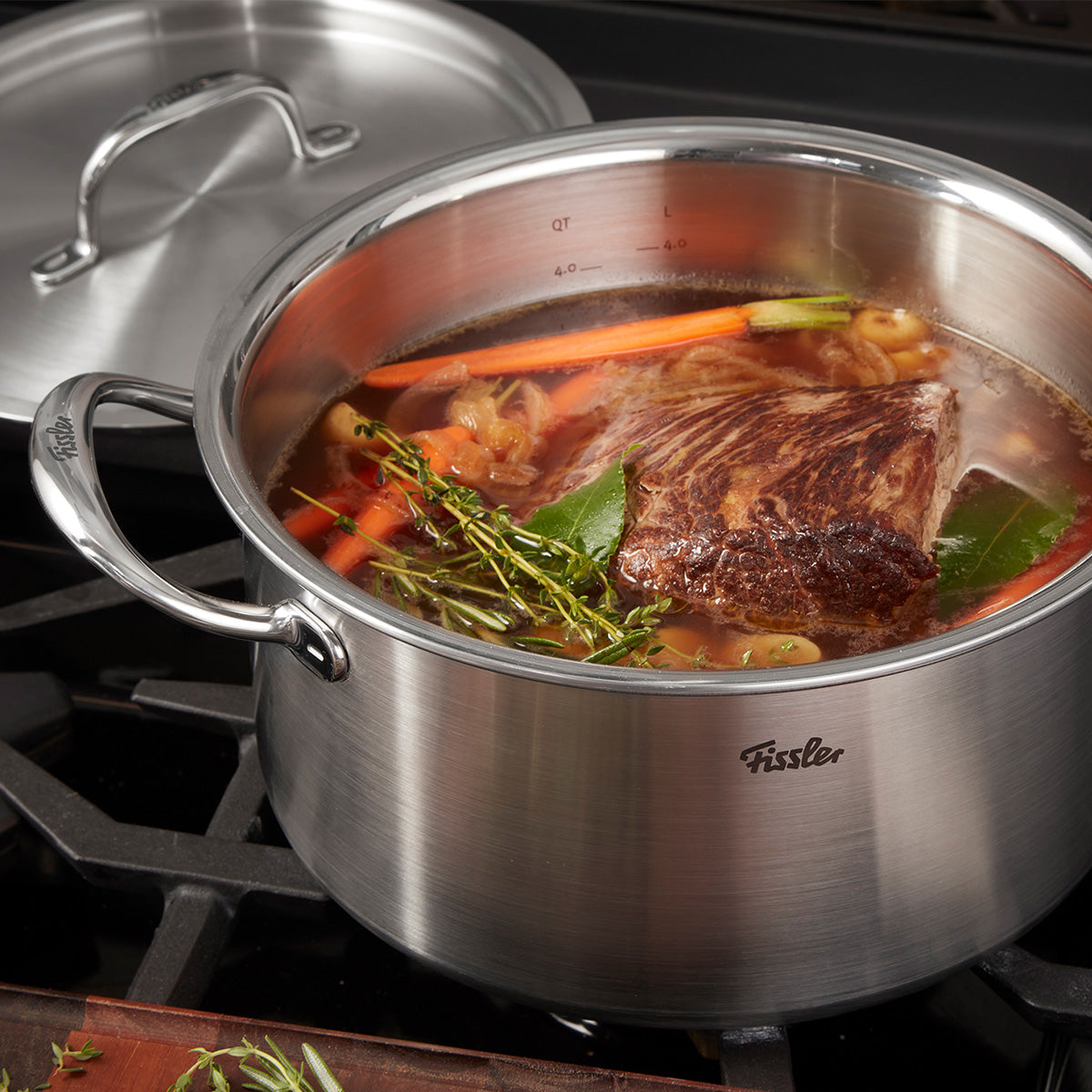 Auf dem Herd steht ein Fissler M5 Pro-Ply®-Kochtopf aus recyceltem Edelstahl mit passendem Metalldeckel. Im Topf köcheln Brühe, frisches Gemüse, Kräuter und ein großzügiges Stück gebratenes Fleisch – perfekt für anspruchsvolles Kochen. Der Topf vereint deutsche Präzision und langlebige Qualität, ist induktionsgeeignet und spülmaschinenfest. Der hochwertige Metalldeckel rundet das zeitlose Design ab und liegt griffbereit daneben.