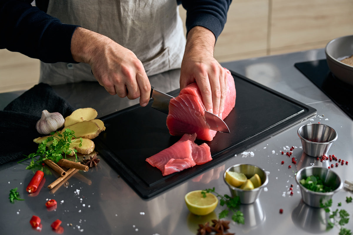 A person demonstrates precision and skill by slicing raw tuna with the Fissler ProCut Santokumesser 17 cm, crafted in Germany and known for its superior cutting performance. The scene features a black cutting board on a sleek stainless steel countertop, accented with fresh herbs, spices, garlic bulbs, ginger root, vibrant chili peppers, and lemon wedges—reflecting the quality culinary experience enabled by Fissler’s expertly engineered kitchen accessories.