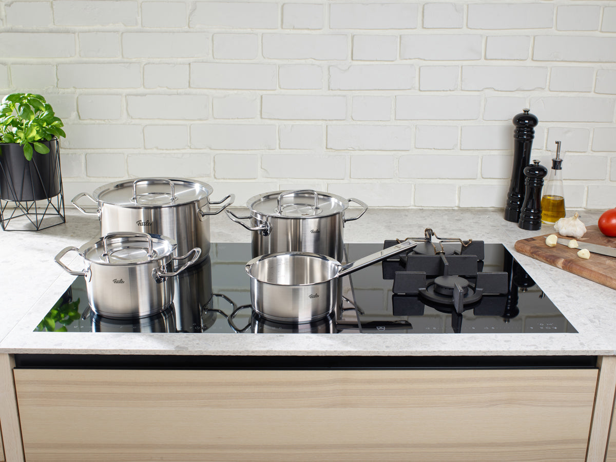 Vier Töpfe der Fissler Original-Profi Collection® mit Metalldeckeln auf einem eleganten Glaskocher, der deutsche Präzision und zeitloses Design zeigt.