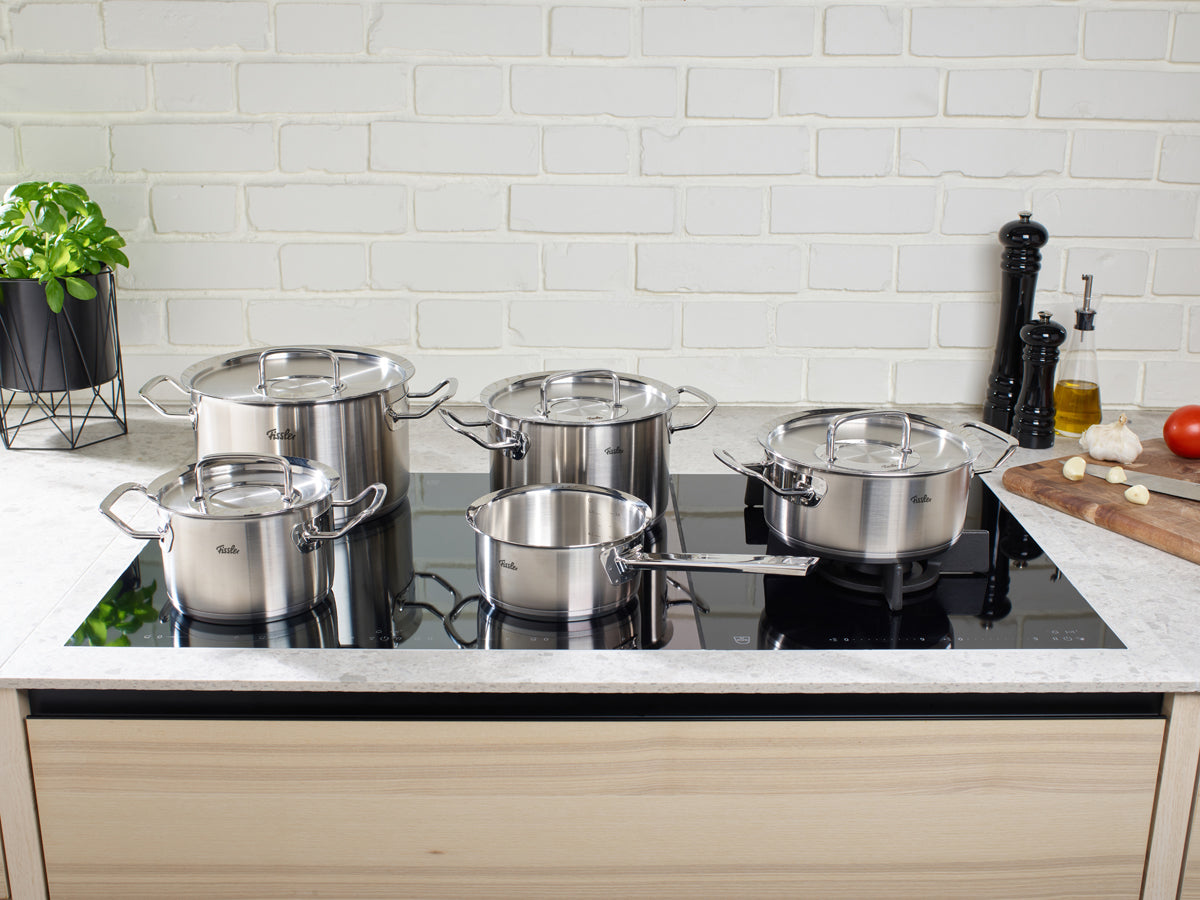 Fünf präzise gefertigte Töpfe und Pfannen aus dem 5-teiligen Set der Fissler Original-Profi Collection® – Kochtöpfe, Bratentopf, Stielkasserolle und Servierpfanne – gefertigt aus hochwertigem 18/10-Edelstahl „Made in Germany“, sind elegant auf einem modernen schwarzen Induktionskochfeld angeordnet. Rechts unterstreicht ein Holzbrett mit geschnittenem Knoblauch, einer Tomate und einer Pfeffermühle vor dem Hintergrund einer klassischen weißen Ziegelwand Fisslers zeitloses Bekenntnis zu Innovation und