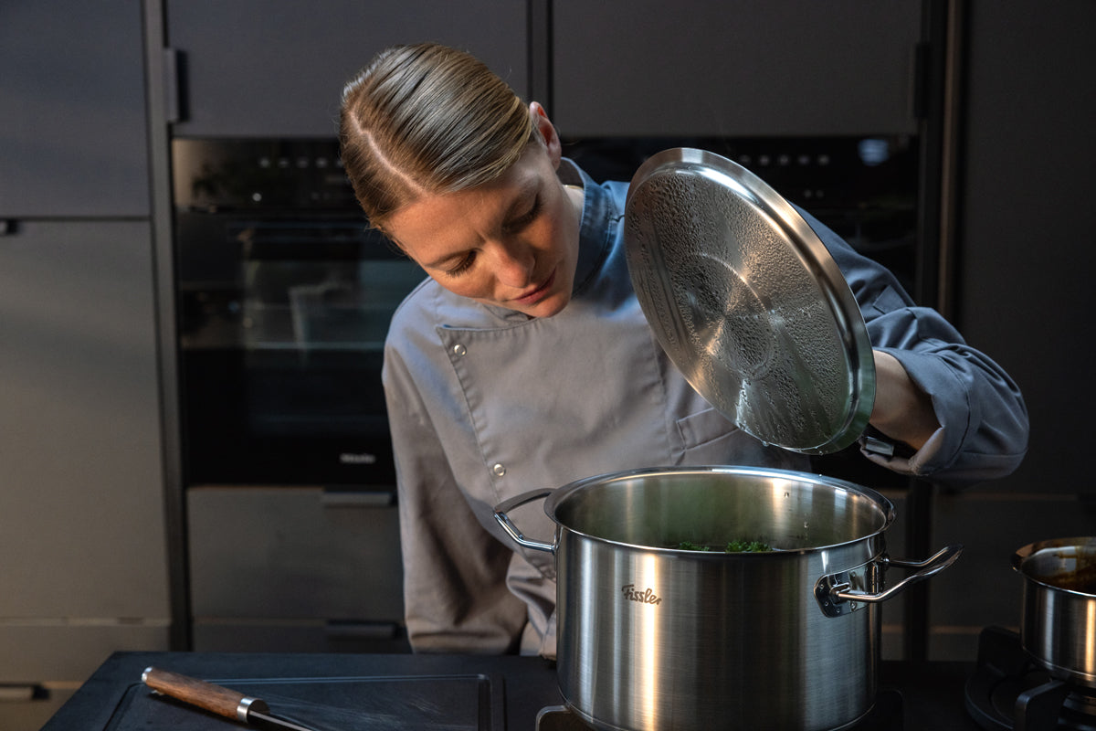 Ein Koch in grauer Uniform hebt den Deckel eines 5-teiligen Edelstahltopf-Sets der Fissler Original-Profi Collection® auf dem Herd an und präsentiert dabei deutsche Präzision und innovatives Design, während er in einer eleganten, modernen Küche eine Mahlzeit zubereitet.