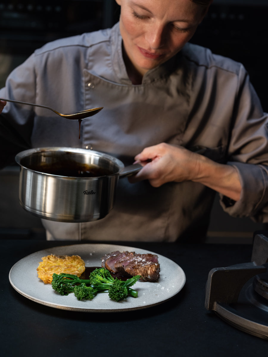 Ein Koch benutzt einen Topf der Fissler Original-Profi Collection®, um Steak, Broccolini und Kartoffeln in einer Profiküche mit Sauce zu übergießen.