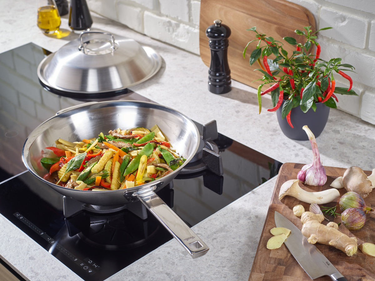 Fissler Original-Profi Collection® Wok 30cm with lid on the stove, stir-frying vegetables; high-quality stainless steel pans and spices nearby.
