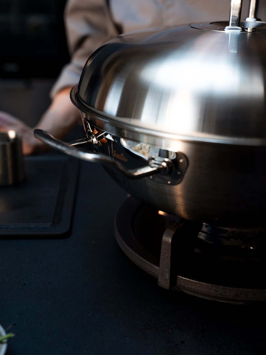 Fissler Original-Profi Collection® Wok 32cm with domed lid, precision-crafted in Germany, on a smooth surface; person behind blurred.