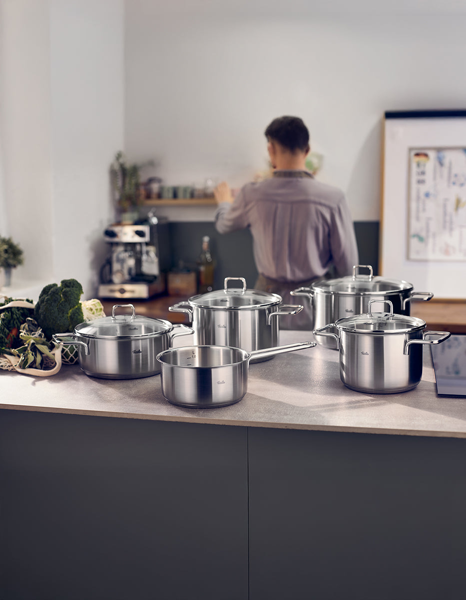 Fissler SatiTherm 5-teiliges Edelstahl-Topfset mit Glasdeckeln auf der Theke, induktionsgeeignet; dahinter frisches Gemüse und Ablagen.