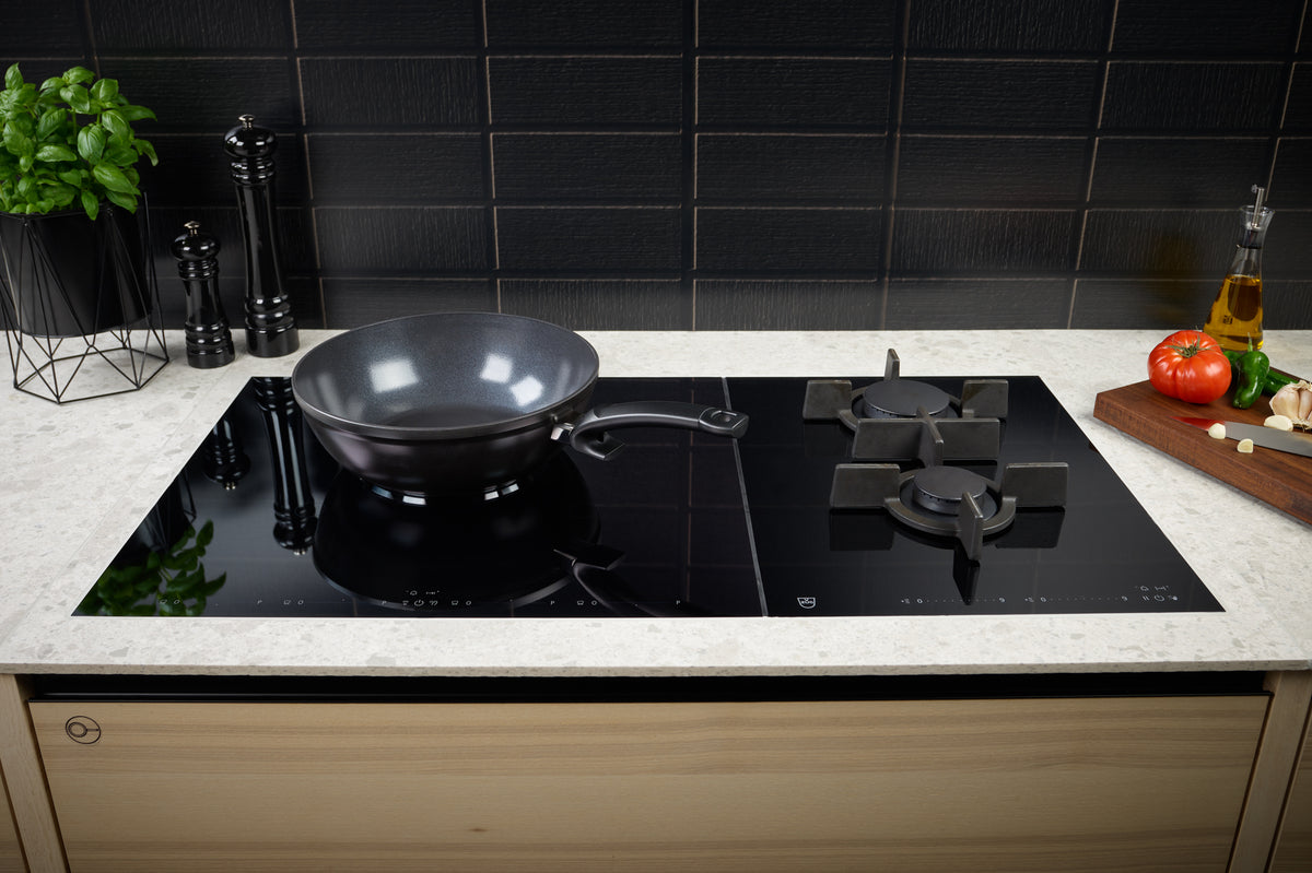 A modern kitchen with the Fissler Ceratal® Wok Orbit Black on a glass ceramic hob, fresh herbs, and ready-to-cook vegetables.