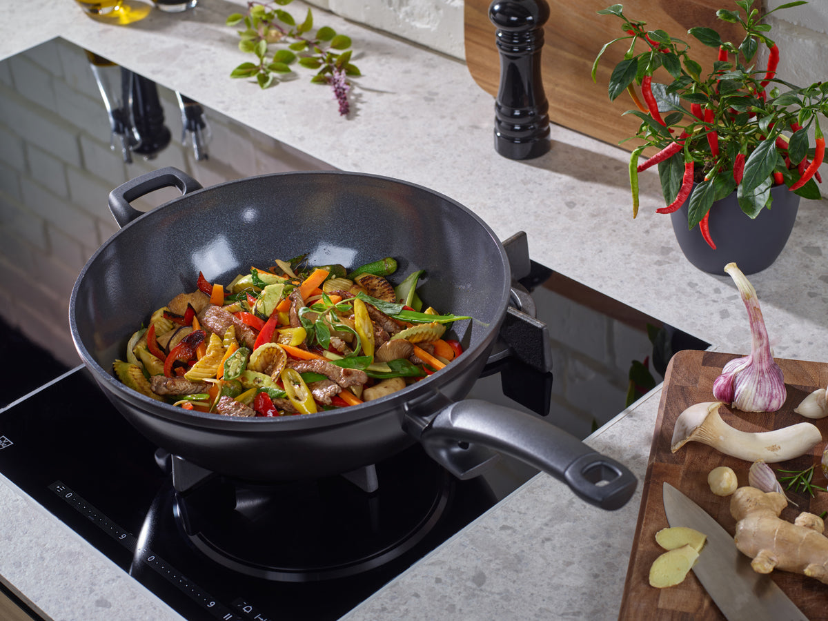 Ceratal® Wok Orbit Black by Fissler, PTFE-free, on the stovetop with stir-fried ingredients; fresh herbs and spices arranged on a modern countertop.