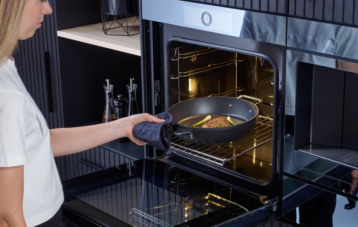 A person uses an oven mitt to place a Fissler Adamant® serving pan with meat and vegetables into a modern built-in oven.