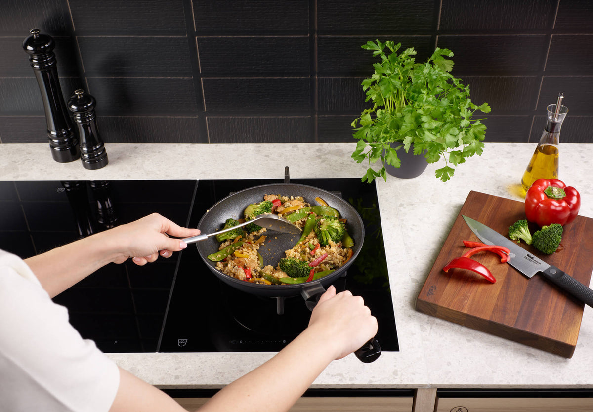 A person cooks with the Fissler Adamant® Classic Frying Pan on the stove, surrounded by fresh herbs, vegetables, and kitchen utensils.