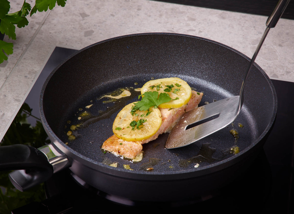Lachsfilet mit Zitrone und Kräutern in einer Fissler Adamant® Comfort Pfanne, die mit einem Spatel angehoben wird; im Hintergrund frische Petersilie.
