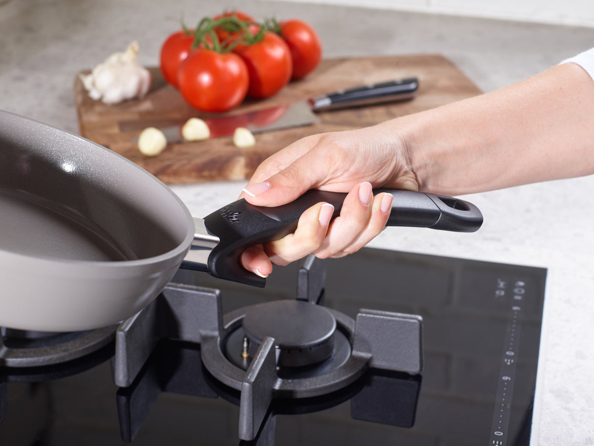 A hand attaches a handle to Fissler Ceratal® Comfort pans on the stove; fresh ingredients and a knife lie on a wooden board next to them.