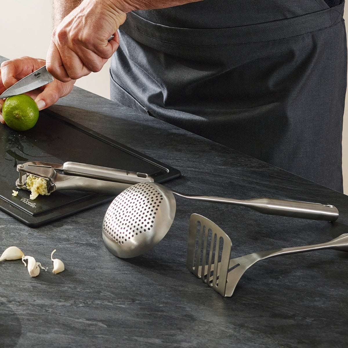 A person in a gray apron prepares food on a modern dark surface next to garlic, a press, and the Fissler Essential® potato masher.