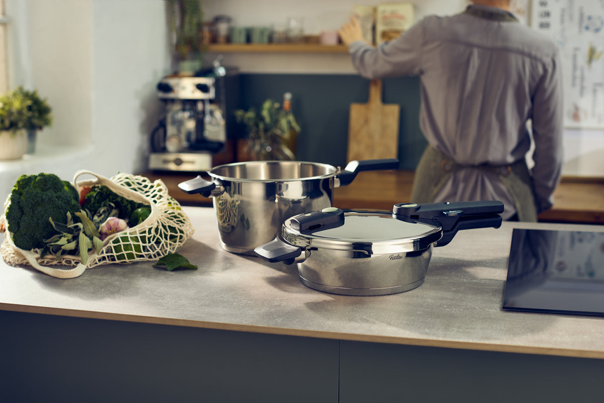 Fissler Vitaquick® Elemental pots (2.5 and 4.5 L) with lids on a kitchen table, vegetables next to them; person in the background.