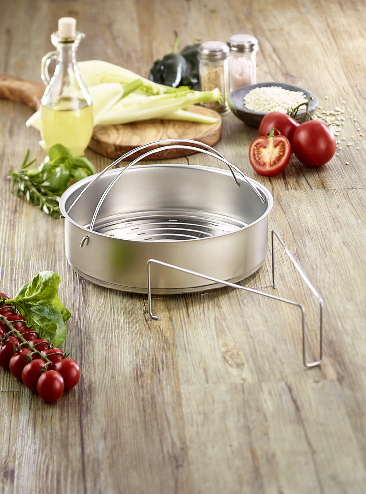 Fissler stainless steel steamer insert with tripod on a wooden surface, surrounded by fresh vegetables and herbs for healthy cooking.