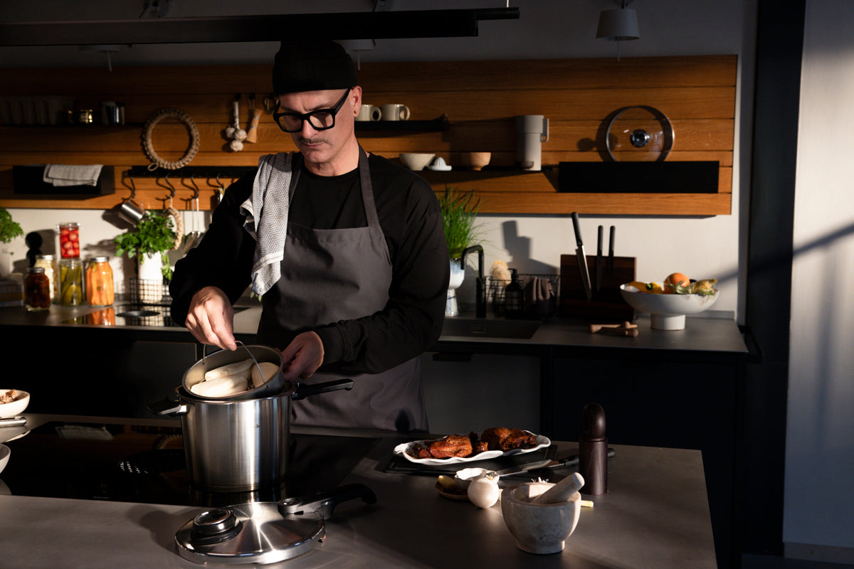In a sunlit modern kitchen, a person with glasses and a beanie expertly stirs a pot on the stove, inspired by fresh Schnellkochtopf Rezepte. Nearby, vibrant ingredients and herbs rest on the counter beside the Fissler Vitavit® Premium 2-tlg. Set mit SKT Buch—a hallmark of German craftsmanship since 1845—ready to deliver precision cooking, durability, and timeless design for another gourmet creation.