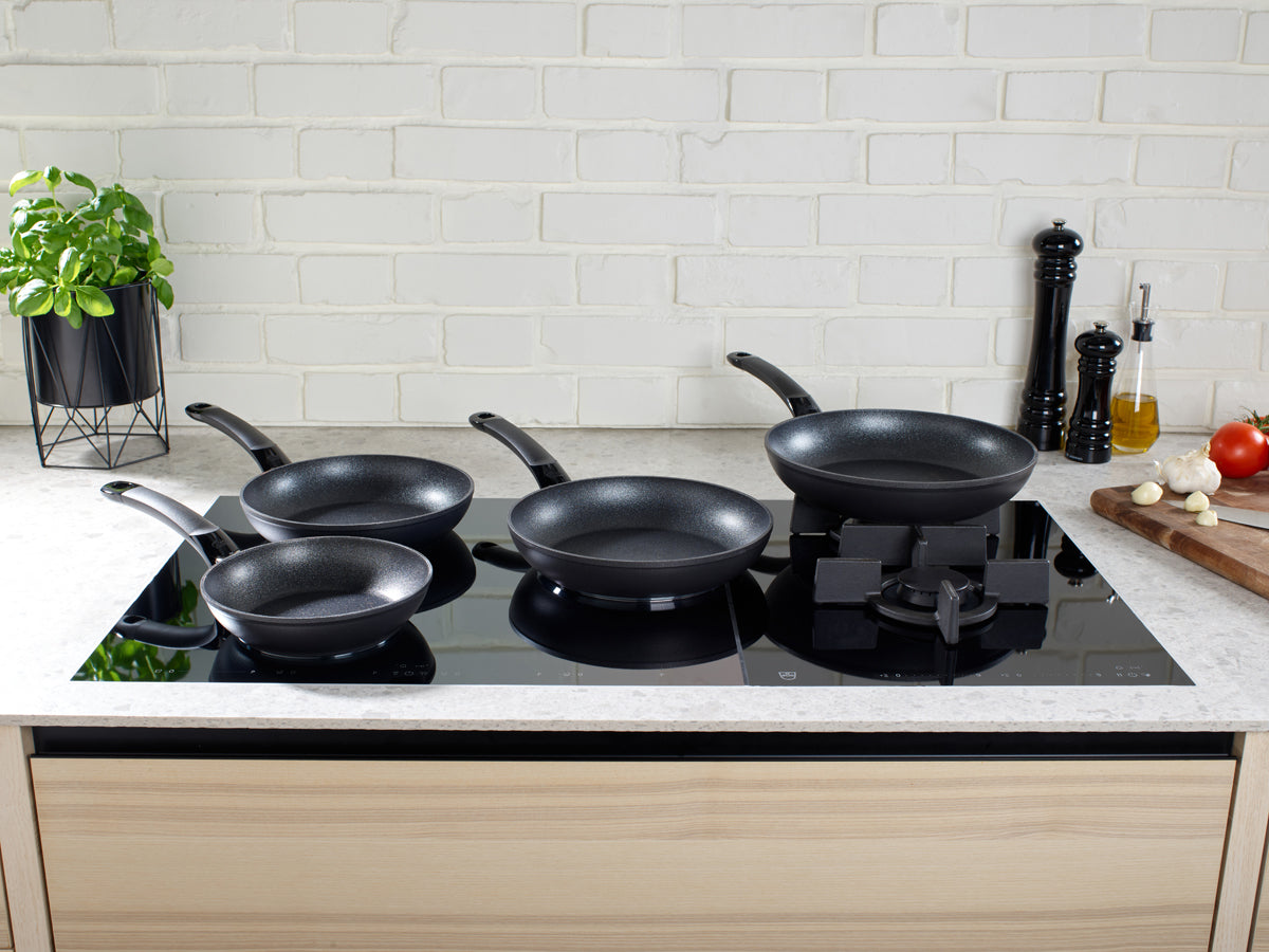 Fünf Fissler Adamant® Classic Pfannen auf einem modernen Herd, der mit Kräutern und frischen Zutaten die Qualität deutscher Herstellung präsentiert.