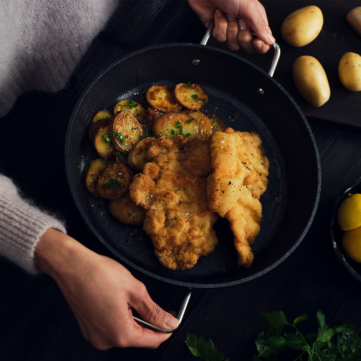 A person showcases the Fissler Adamant® Servierpfanne, featuring two perfectly breaded, fried schnitzels and golden sautéed potato slices artfully garnished with herbs. The innovative anti-stick coating and precision German engineering—hallmarks of Fissler’s heritage since 1845—deliver effortless performance in every meal. Nearby, whole potatoes and fresh parsley rest on a sophisticated dark tabletop, highlighting the premium quality and timeless design of authentic German-made cookware.