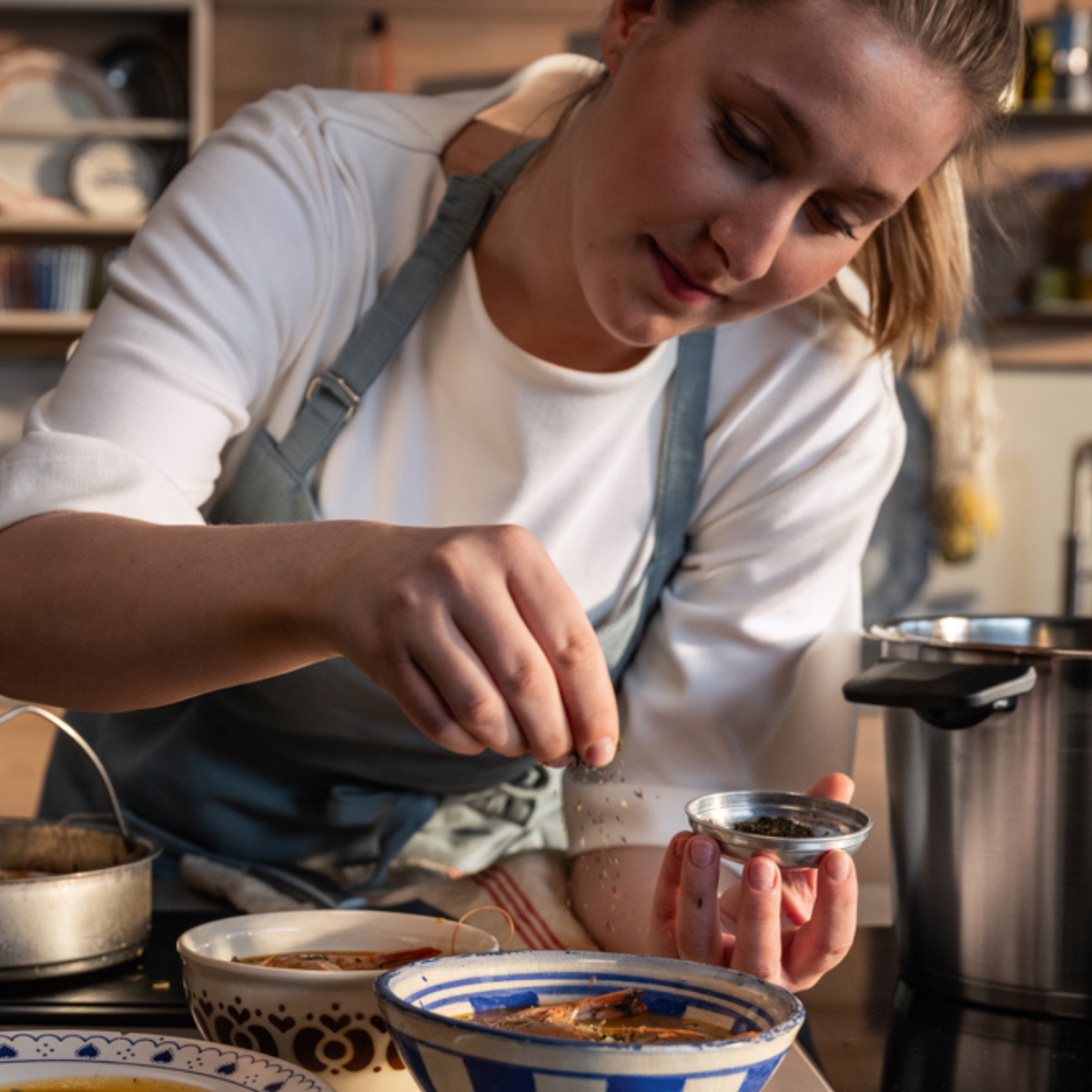 Eine Person in einer Schürze streut in einer Küche Gewürze über eine Schüssel mit Essen, umgeben von hochwertigen Fissler-Edelstahltöpfen und -Kochgeschirr. Die Szene spiegelt Fisslers Tradition von Präzisionstechnik, zeitlosem Design und Innovation „Made in Germany“ wider.