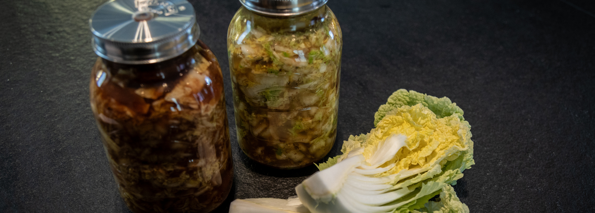 Two glass jars filled with homemade kimchi are displayed on a dark countertop beside a halved head of fresh napa cabbage, evoking precision and freshness. The setting highlights the importance of quality kitchen tools—such as Fissler’s stainless steel cookware or accessories—in preparing and storing delicious homemade creations.