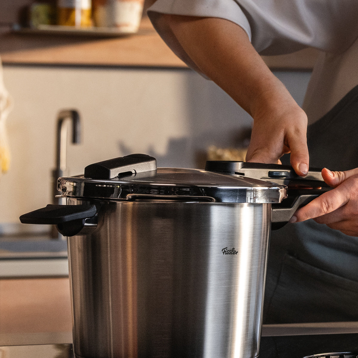 Eine Person schließt einen Fissler Schnellkochtopf aus rostfreiem Stahl auf einer Arbeitsplatte in einer warm beleuchteten Küche, deutsche Qualität ist überall sichtbar.