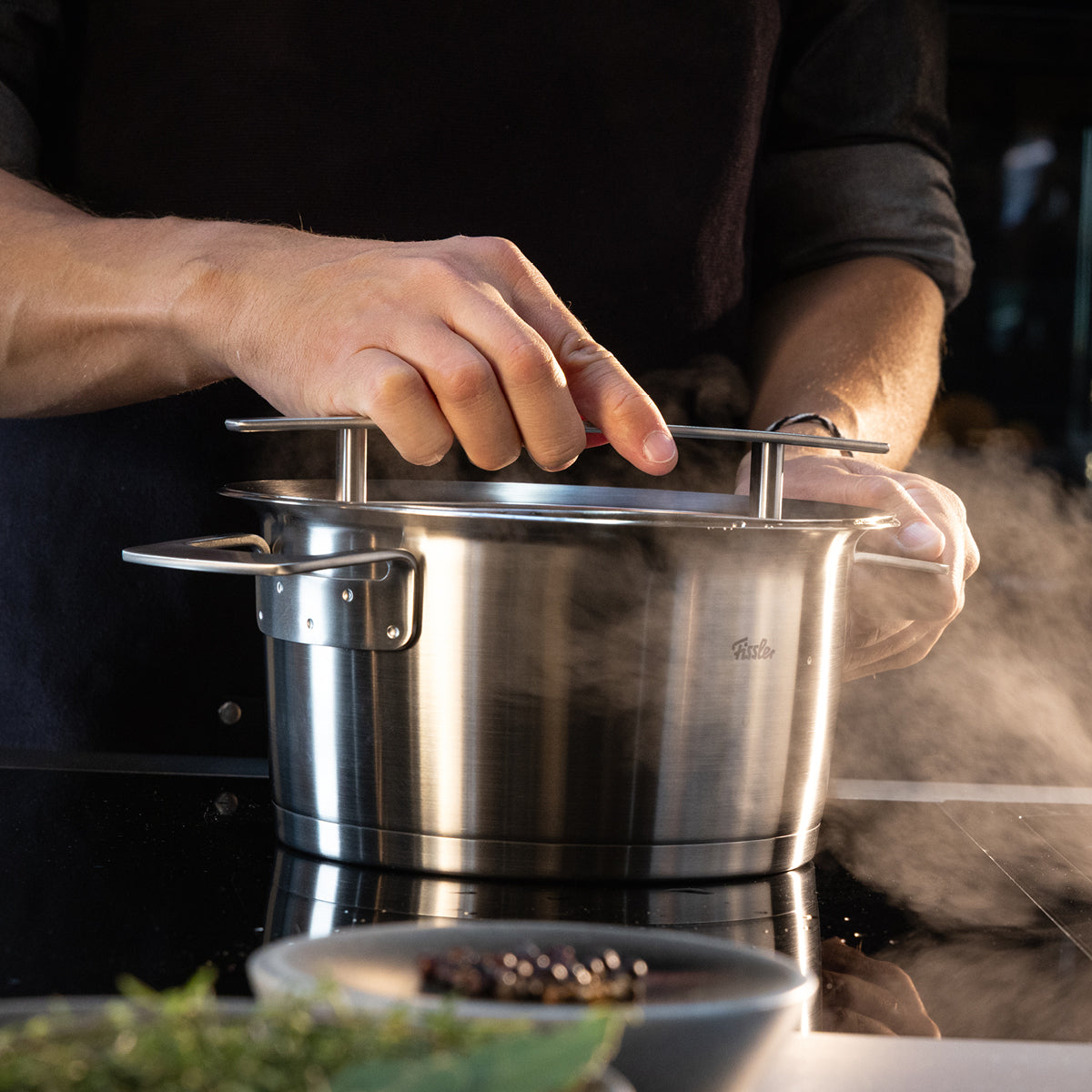 Die Hände heben den Deckel von einem dampfenden Fissler-Edelstahltopf auf dem Herd; im Vordergrund liegen frische Kräuter.