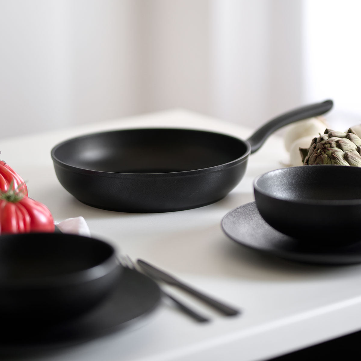 Fissler Cenit® induction frying pan with non-stick coating on a white table, next to kitchen utensils and fresh vegetables in soft light.