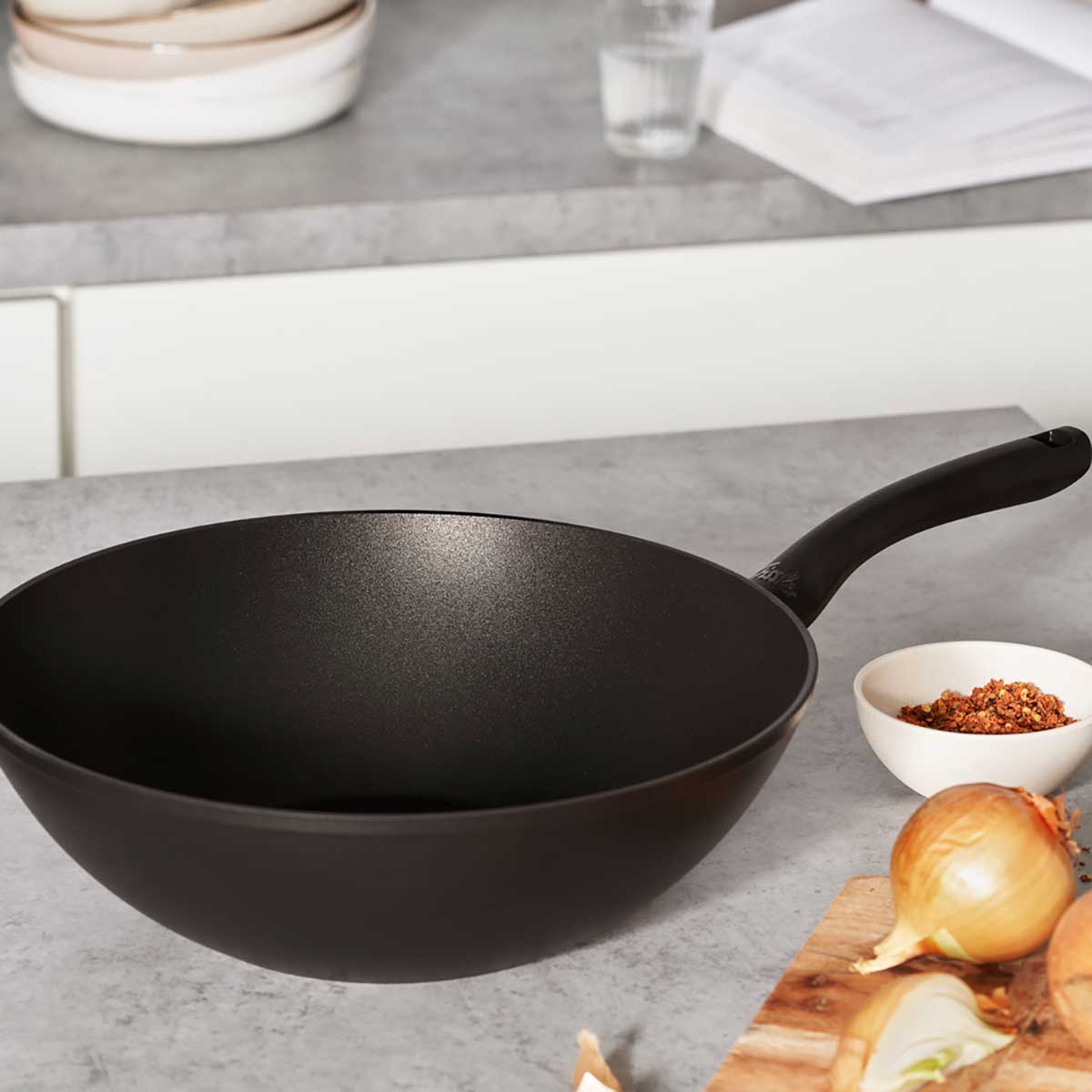 Die Fissler Cenit® Wokpfanne Induktion 28 cm - Präzision und Design. Umgeben von Gewürzen, Wasser, Kochbuch und Schneidebrett.