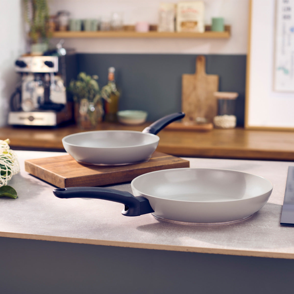 Erleben Sie deutsche Präzision und zeitloses Design mit zwei Fissler Ceratal® Evo Classic-Pfannen mit ergonomischen schwarzen Griffen, die elegant auf einer Küchenarbeitsplatte präsentiert werden – eine davon kunstvoll auf einem Holzschneidebrett platziert. Diese hochwertigen Pfannen sind mit einer innovativen Antihaft-Keramikbeschichtung für müheloses Kochen und einfache Reinigung ausgestattet und verkörpern Fisslers Tradition von Langlebigkeit und technischer Exzellenz seit 1845. Im Hintergrund entdecken