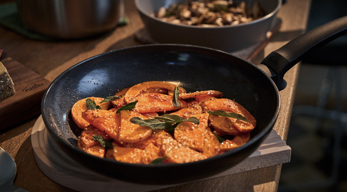 Frisch angebratenes orangefarbenes Gemüse wie Süßkartoffeln oder Kürbis, elegant mit Kräutern garniert und in einer hochwertigen schwarzen Fissler-Pfanne auf einem Holztisch serviert. Im Hintergrund unterstreicht eine Schale mit Pilzen die Vielseitigkeit des hochwertigen Kochgeschirrs und das zeitlose deutsche Design.