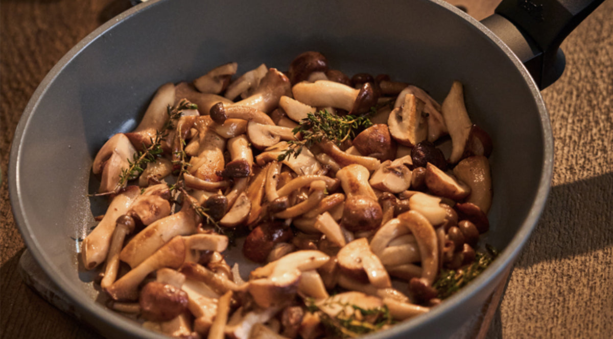 In einer hochwertigen Fissler-Pfanne aus erstklassigem Edelstahl werden perfekt angebratene Pilzmischungen präsentiert, garniert mit frischen Kräuterzweigen. Die Pfanne steht elegant auf einer Holzarbeitsplatte und verkörpert deutsche Präzisionstechnik und zeitloses Design.