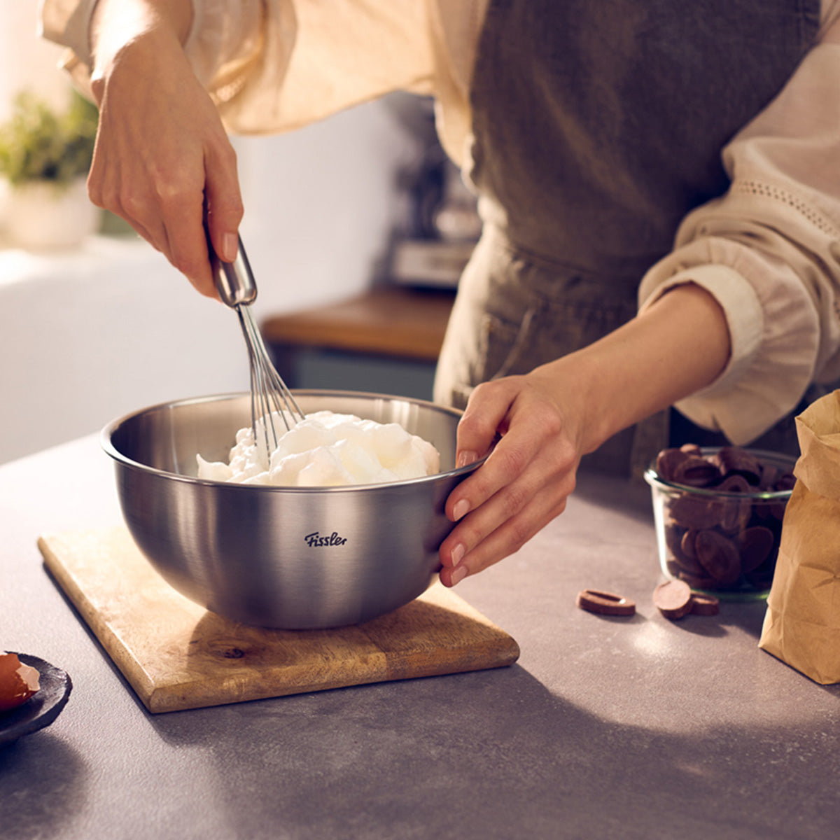 A person stirs ingredients in a Fissler Essential® 20cm bowl; chocolate and a paper bag sit on a premium kitchen counter.