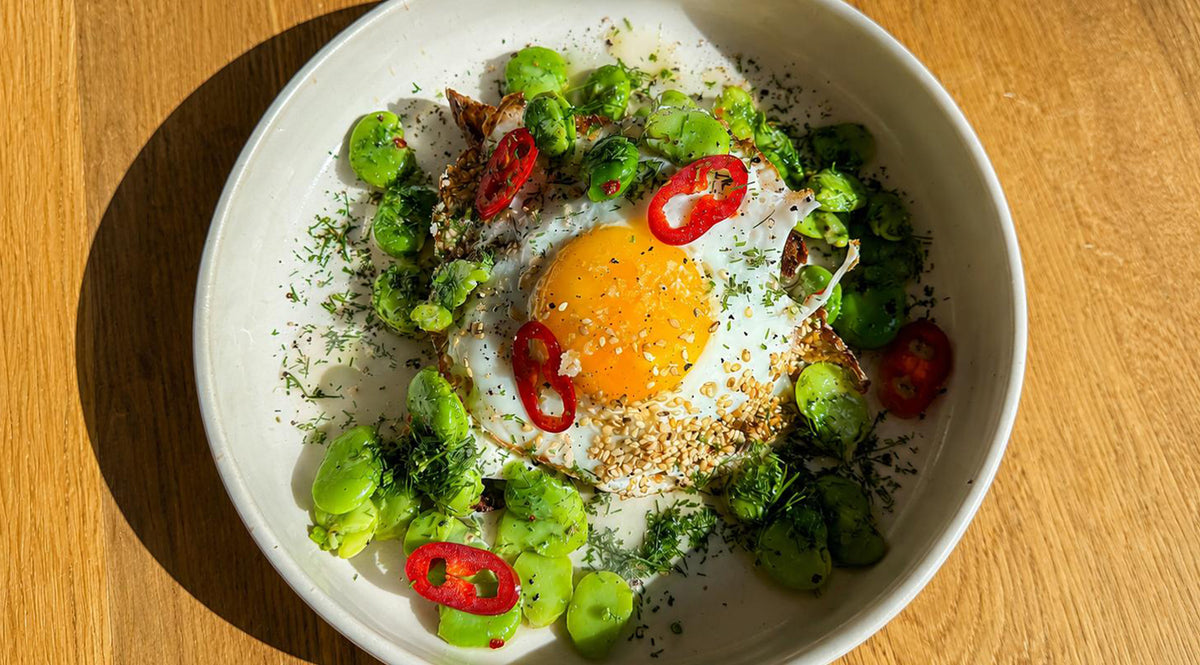 Auf einem Holztisch steht eine wunderschön angerichtete weiße Schüssel mit einem Spiegelei, Saubohnen, roten Chilischeiben, frischen Kräutern und Samen. Das Gericht wird im zeitlosen Stil zubereitet und präsentiert, der für Fisslers präzisionsgefertigtes Kochgeschirr charakteristisch ist und in jedem Detail Langlebigkeit und Innovation widerspiegelt.