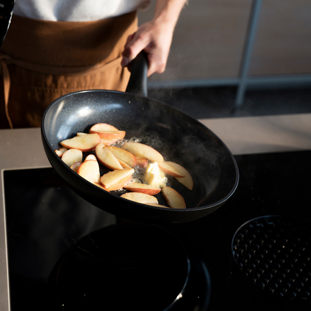 Sliced apples caramelize in a Fissler Essential® Frying Pan Set, steam rises as the cook’s arm and upper body show Made in Germany precision.