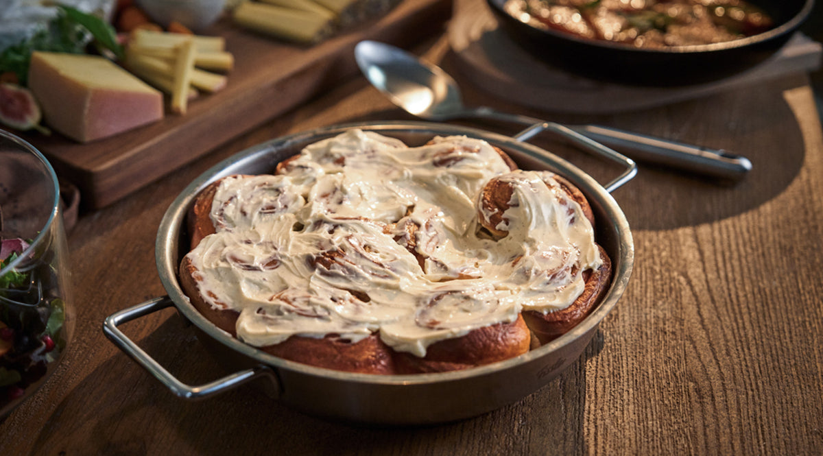Eine wunderschön gearbeitete Fissler-Edelstahlpfanne präsentiert warme Zimtschnecken mit cremiger weißer Glasur, die auf einem Holztisch neben Käse, Obst und einem Löffel im Hintergrund angerichtet sind – ein Beispiel für Fisslers Tradition der Präzisionstechnik und des zeitlosen deutschen Designs.