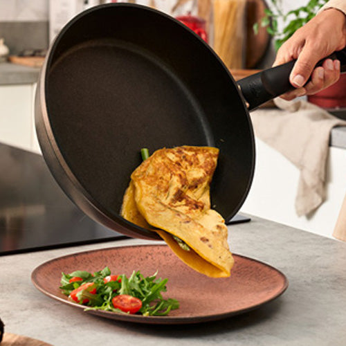 Eine Hand kippt die Fissler Levital®+ Comfort Pan und serviert ein Omelett auf einem Teller - Präzision, Langlebigkeit und Qualität Made in Germany.