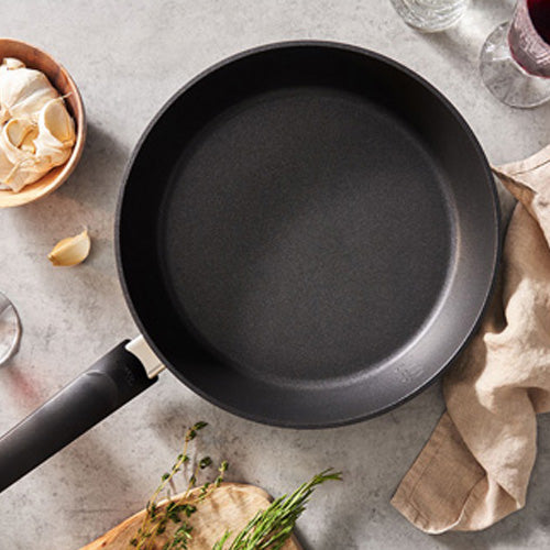 Fissler Levital®+ Comfort Pfanne auf grauer Arbeitsplatte mit beigem Tuch, Knoblauch, Kräutern und Gläsern - zeitloses Präzisionskochgeschirr aus deutscher Produktion.