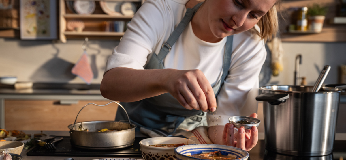 A person in an apron prepares food, sprinkling herbs into a bowl surrounded by Fissler cookware and kitchen accessories.