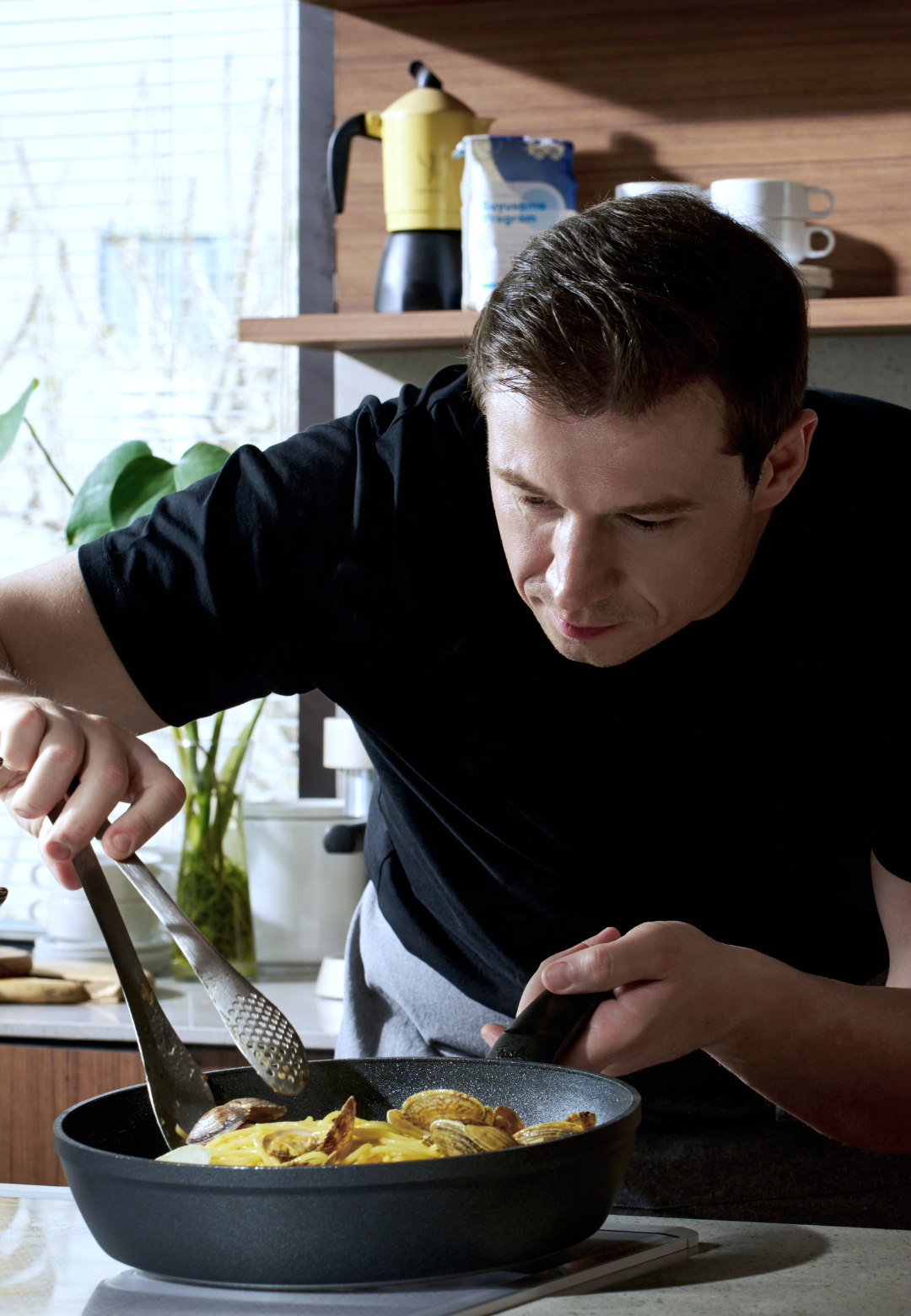 Ein Mann in einem schwarzen Hemd bereitet in einer hochwertigen Fissler-Edelstahlpfanne Essen zu und geht dabei gekonnt mit einer Zange und einem Pfannenwender um. Die helle Küche wird durch üppige Pflanzen und modernes Fissler-Kochgeschirr sowie Zubehör, das im Hintergrund zu sehen ist, akzentuiert.