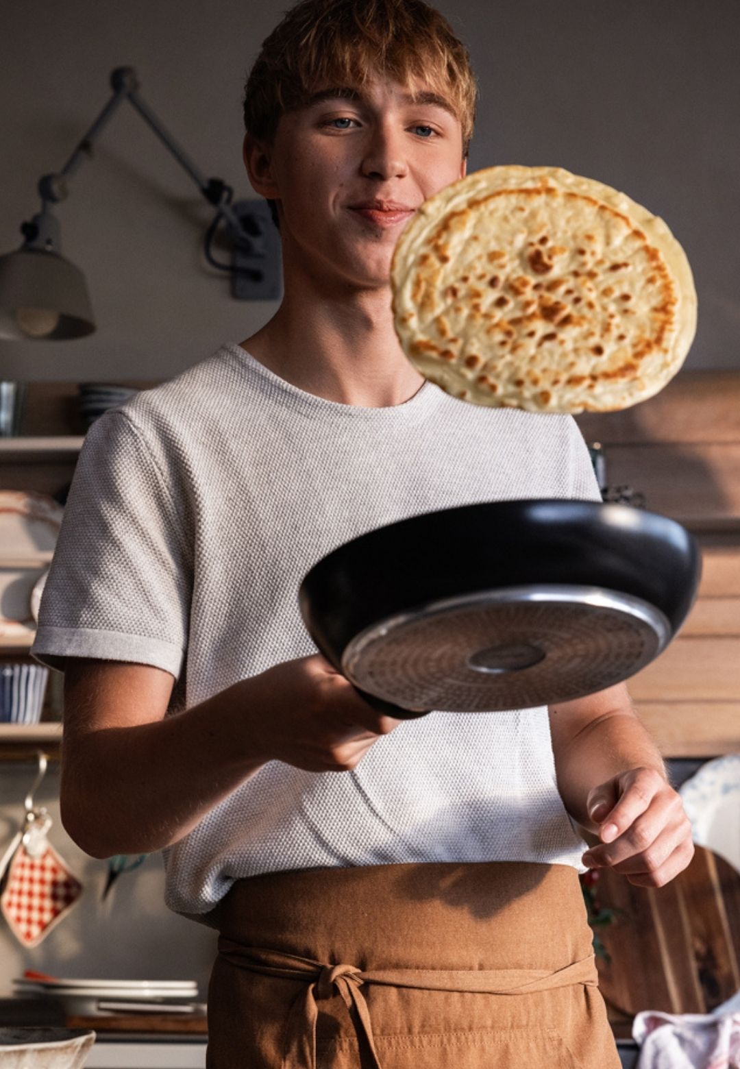 A young person flips a pancake in a black Fissler frying pan, showcasing German quality and timeless design in a cozy kitchen.