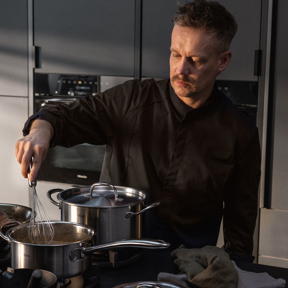 In einer modernen Küche rührt eine Person in einer dunklen Kochjacke gekonnt mit einem Schneebesen im Fissler M5 Pro-Ply® Kochtopf mit Metalldeckel um, der fachmännisch aus recyceltem Edelstahl gefertigt und für optimale Leistung auf einem Induktionsherd ausgelegt ist. Daneben steht ein weiterer hochwertiger Fissler-Topf. Beide hochwertigen Töpfe verkörpern die seit 1845 bestehende Tradition von Fissler in Sachen deutscher Präzision, Langlebigkeit und zeitlosem Design und sind spülmaschinenfest, was die